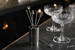 A realistic premium bar scene showing custom cocktail stirrers as branded metal drink accessories.